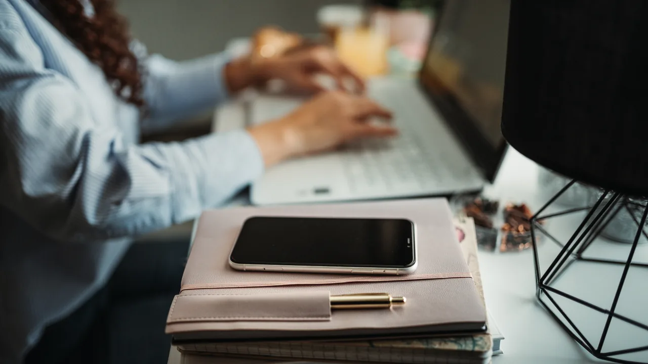Pessoa trabalhando no laptop ao lado de um smartphone em uma mesa de escritório