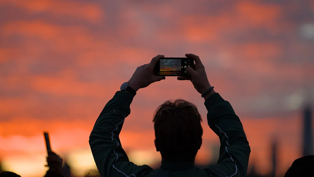 Pessoa fotografando o pôr do sol com um celular; a imagem representa o compartilhamento de fotos e seus dados associados.