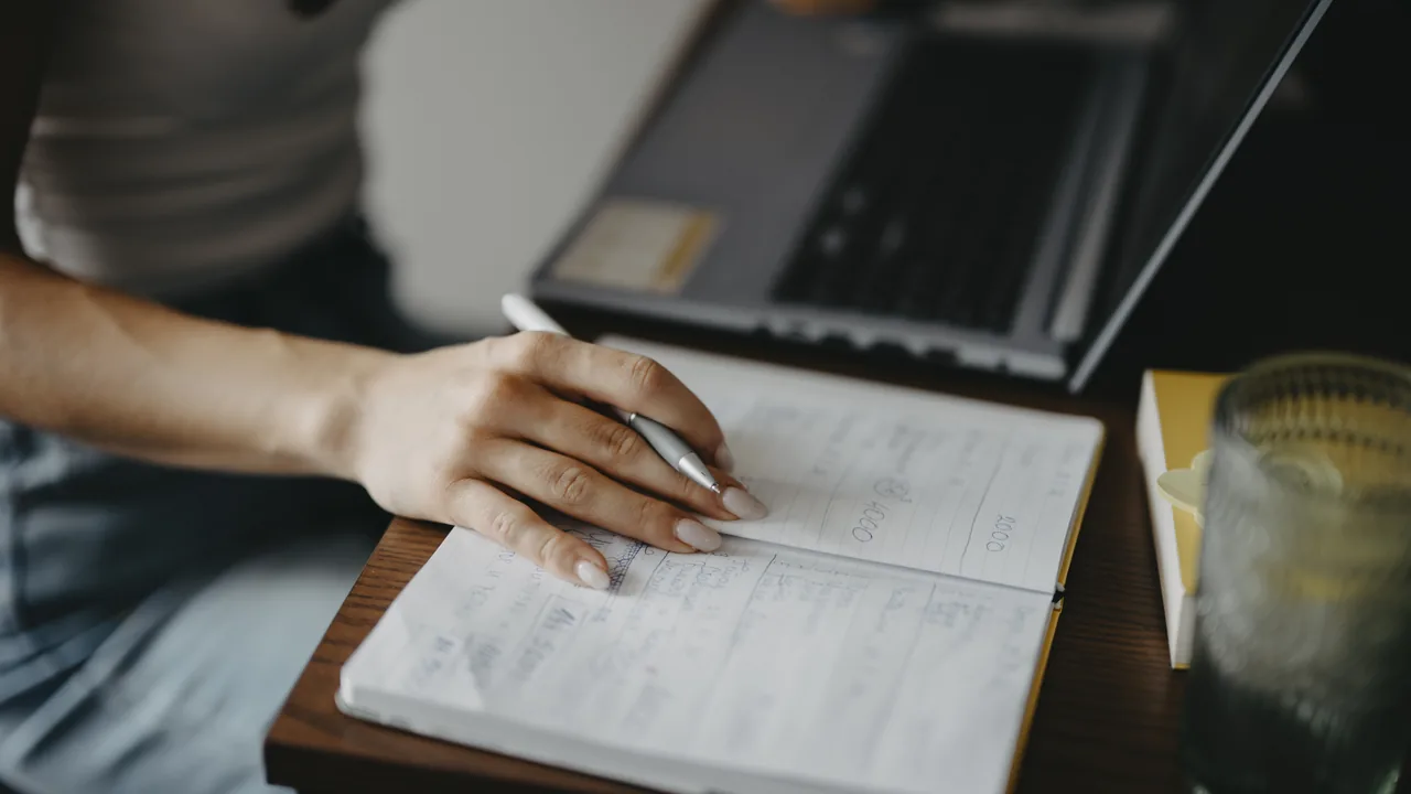 Pessoa fazendo anotações ao lado de um notebook em uma mesa