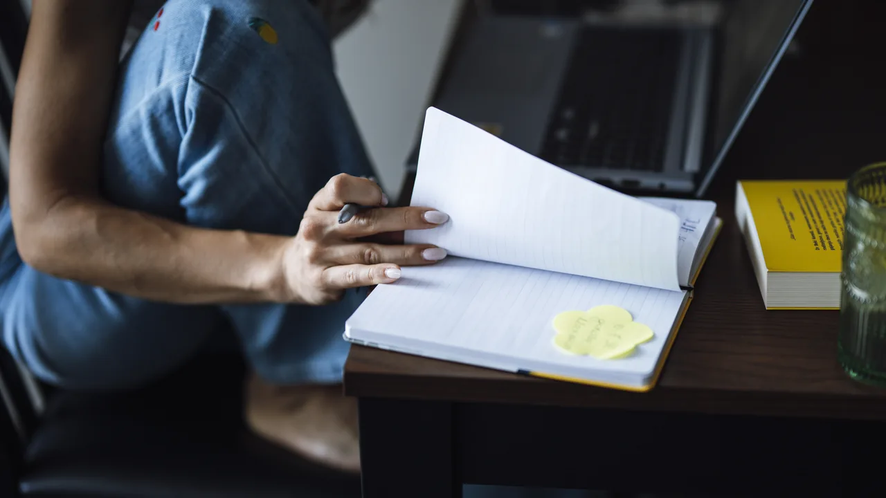 Pessoa escrevendo em um caderno ao lado de um laptop, uma cena que representa a definição de categorias para uma lista