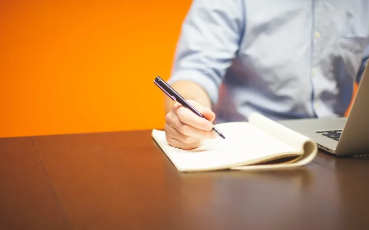Pessoa escrevendo ao lado de um notebook, imagem ilustrativa para assinatura profissional de e-mail