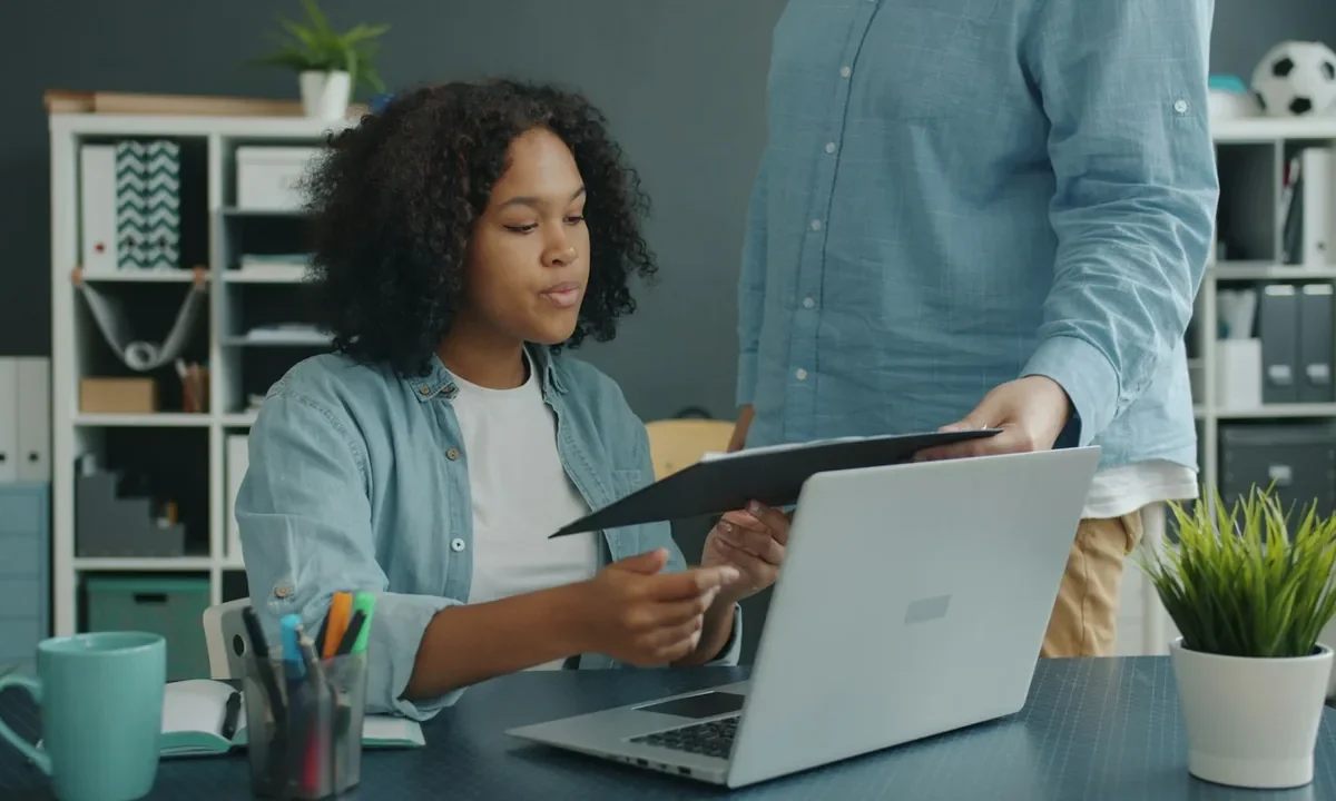 Duas pessoas organizam documentos ao lado de um notebook em uma mesa