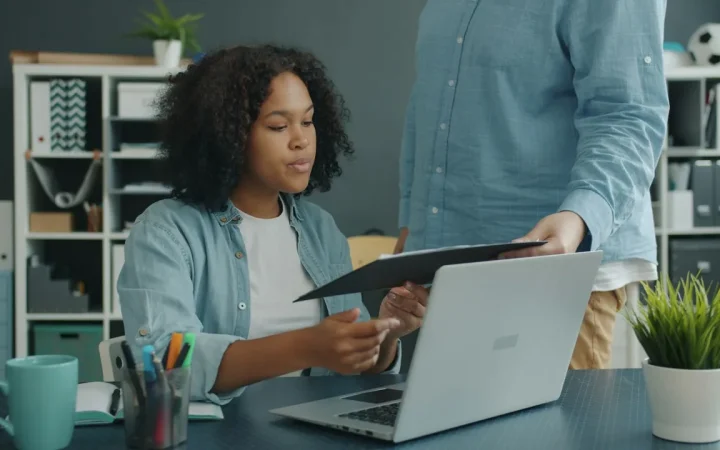 Duas pessoas organizam documentos ao lado de um notebook em uma mesa